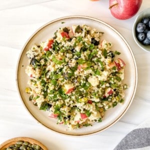 apple quinoa salad on a brown plate next to a red apple and bowl of blueberries.