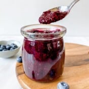 blueberry chia jam in a glass jar on a wooden board next to a bowl of blueberries.