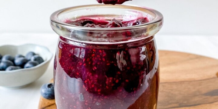 blueberry chia jam in a glass jar on a wooden board next to a bowl of blueberries.