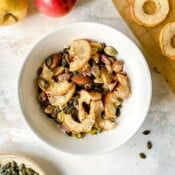 apple trail mix in a white bowl next to red apples and seeds.