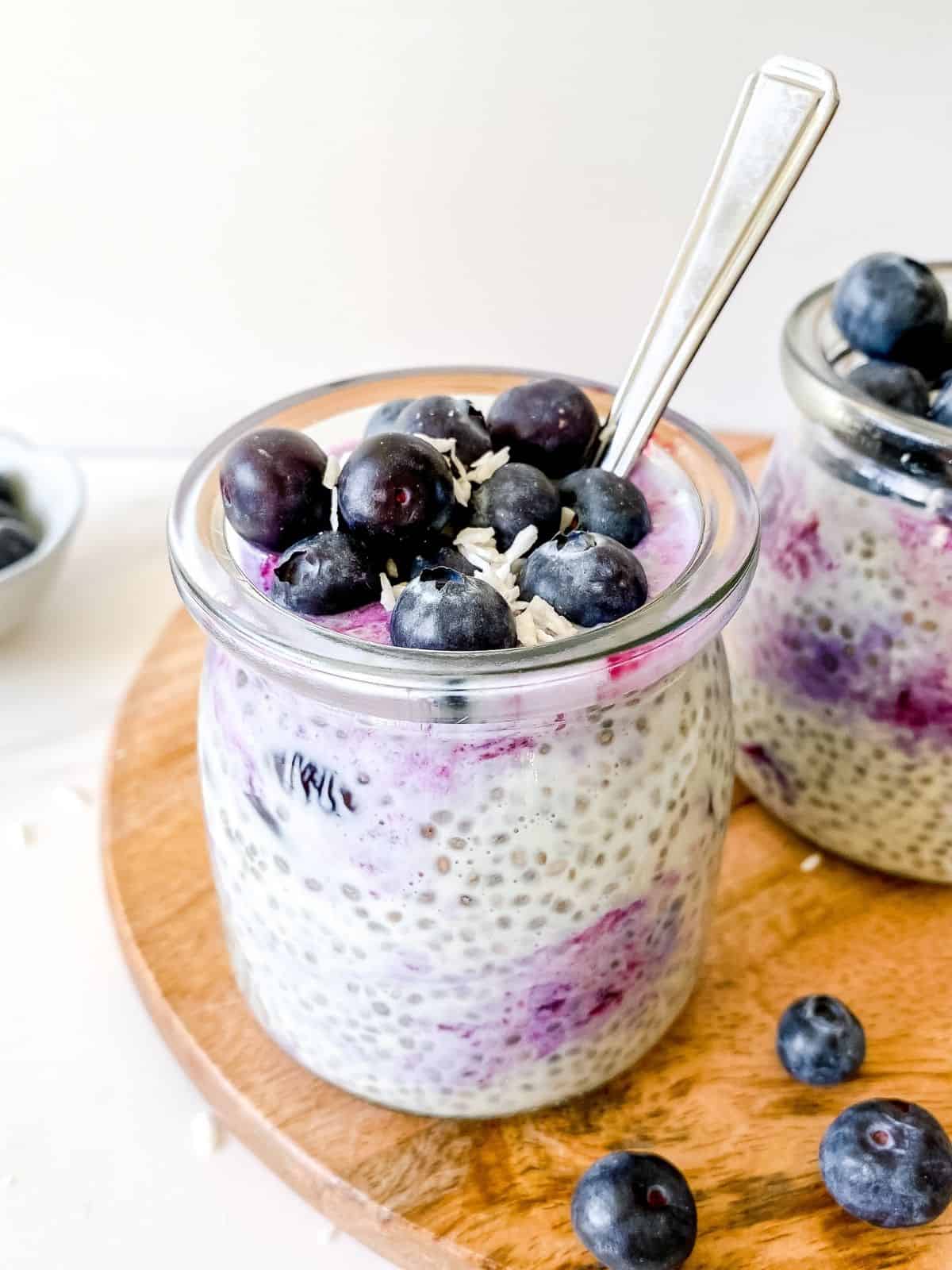 blueberry coconut chia pudding in two glass jars on a wooden board next to blueberries.