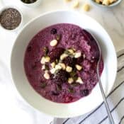 berry quinoa porridge in a white bowl with a spoon on a tea towel next to small bowls of nuts and seeds.