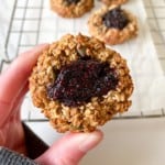 person holding a blueberry thumbprint cookie with a wire rack of cookies in the background.