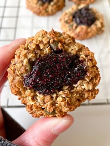 person holding a blueberry thumbprint cookie with a wire rack of cookies in the background.