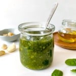 jar of macadamia nut pesto with a spoon in it next to garlic, basil leaves and a bottle of olive oil.