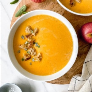 two bowls of butternut squash and apple soup on a round wooden board.