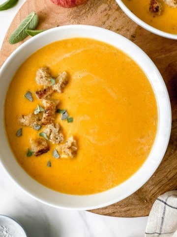 two bowls of butternut squash and apple soup on a round wooden board.