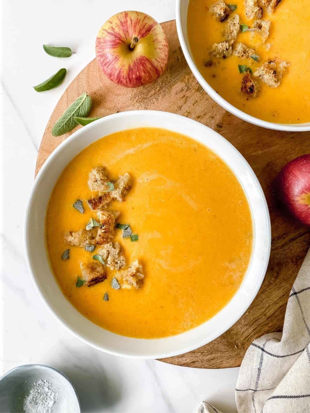 butternut squash and apple soup in two white bowls on a board.