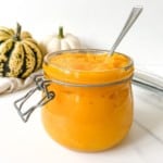 glass jar of pumpkin puree with a spoon in it next to white and yellow pumpkins.