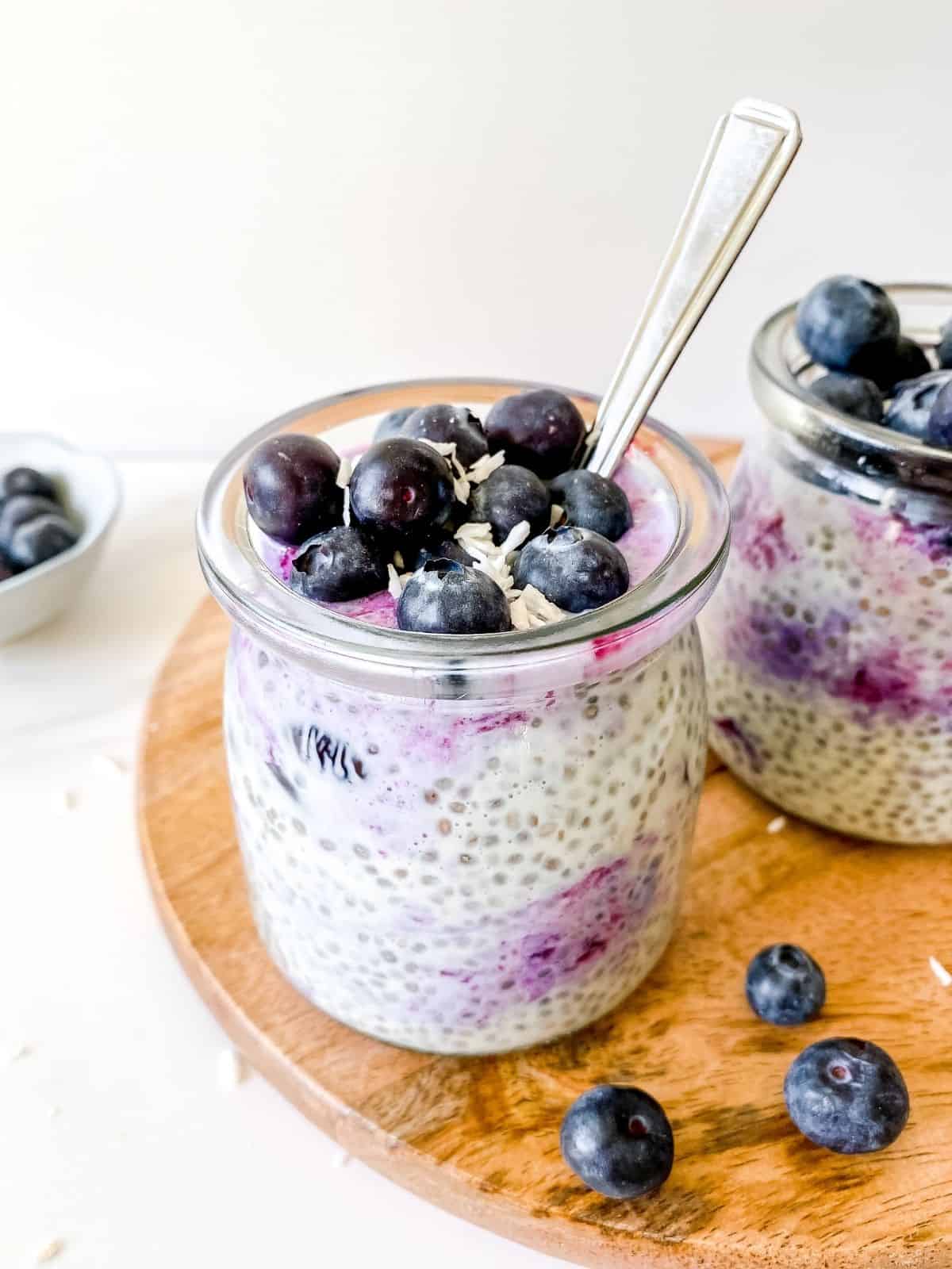 blueberry coconut chia pudding in two glass jars with spoons in them on a wooden board next to a bowl of blueberries.