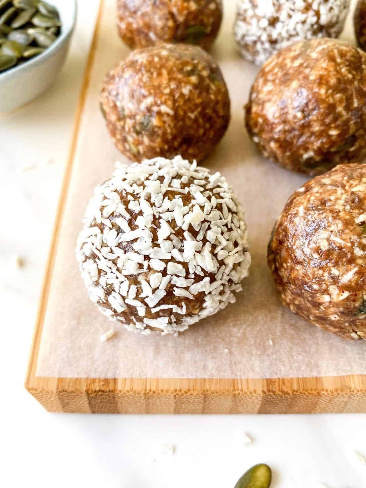 coconut energy balls on a wooden chopping board next to a bowl of pumpkin seeds.