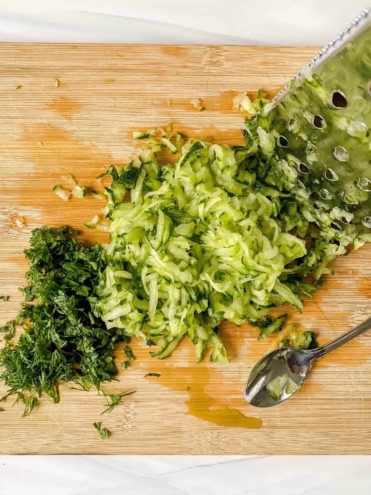 grated cucumber and diced mint and dill on a wooden chopping board.