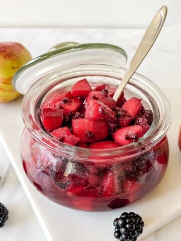apple and blackberry compote in a glass jar with a spoon in it.