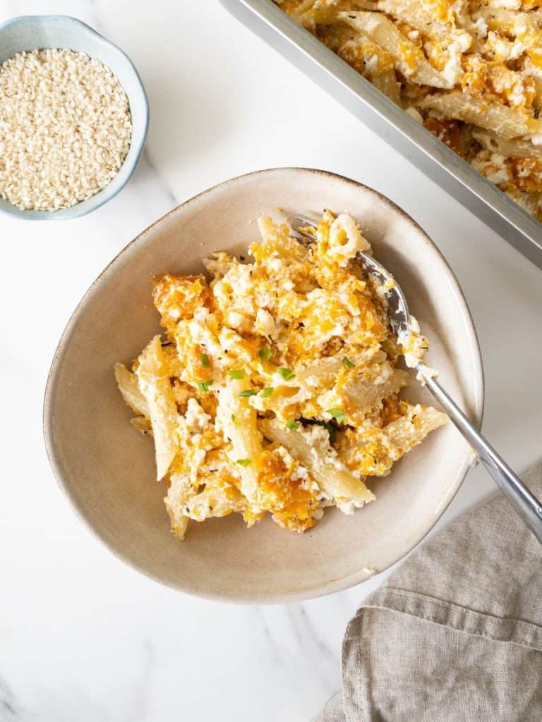 baked ricotta and butternut squash pasta in a light cream bowl next to a baking tray of pasta and a bowl of sesame seeds.