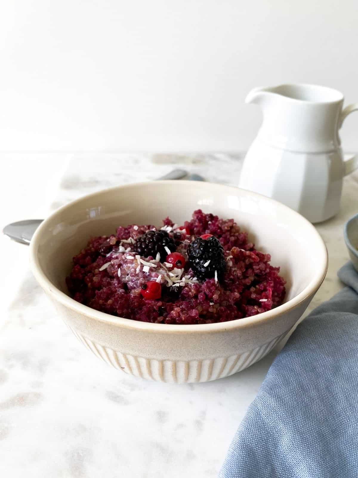 side view of baked quinoa breakfast with blackberries in a cream bowl with a jug in the background.