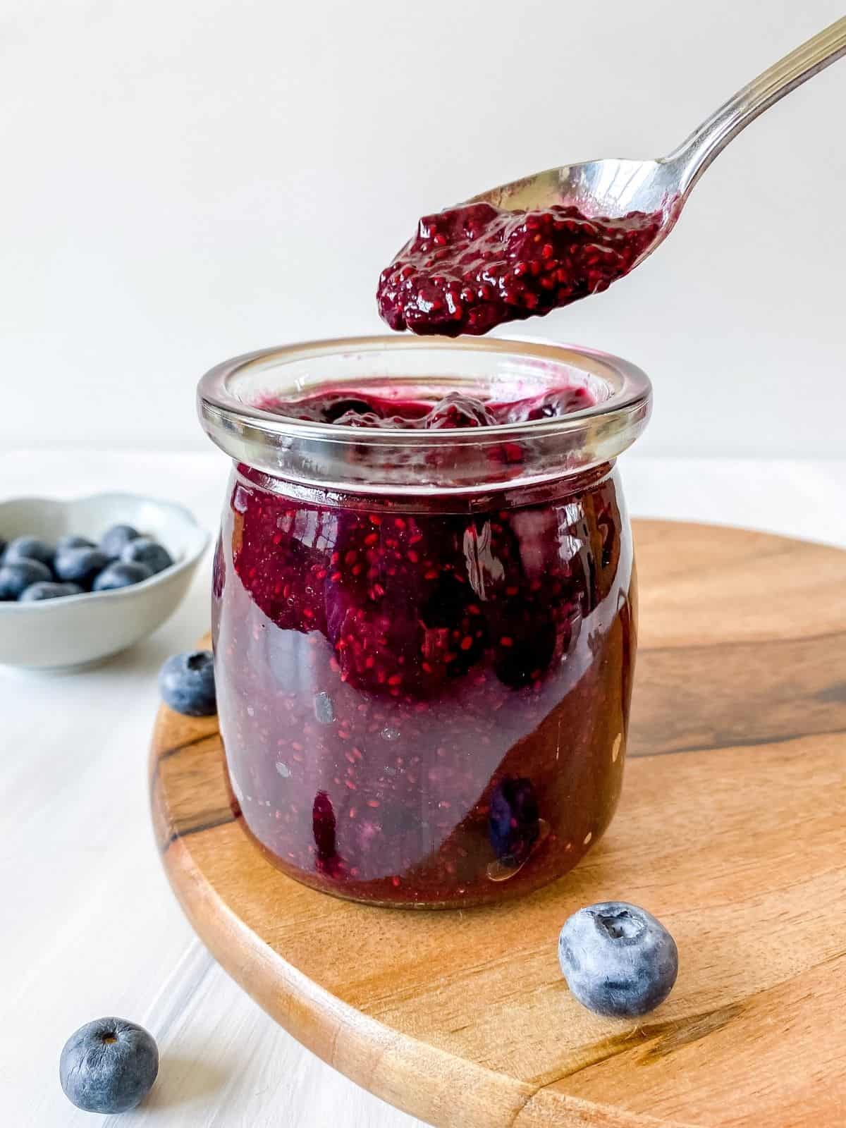 blueberry chia jam in a glass jar on a wooden board with a spoon scooping jam out of it.
