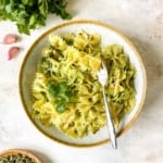 coriander pesto pasta in a light grey bowl with a fork next to a bunch of coriander and bowl of pumpkin seeds.