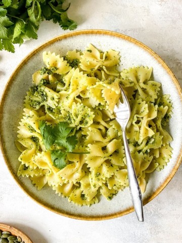 coriander pesto pasta in a light grey bowl with a fork next to a bunch of coriander and bowl of pumpkin seeds.