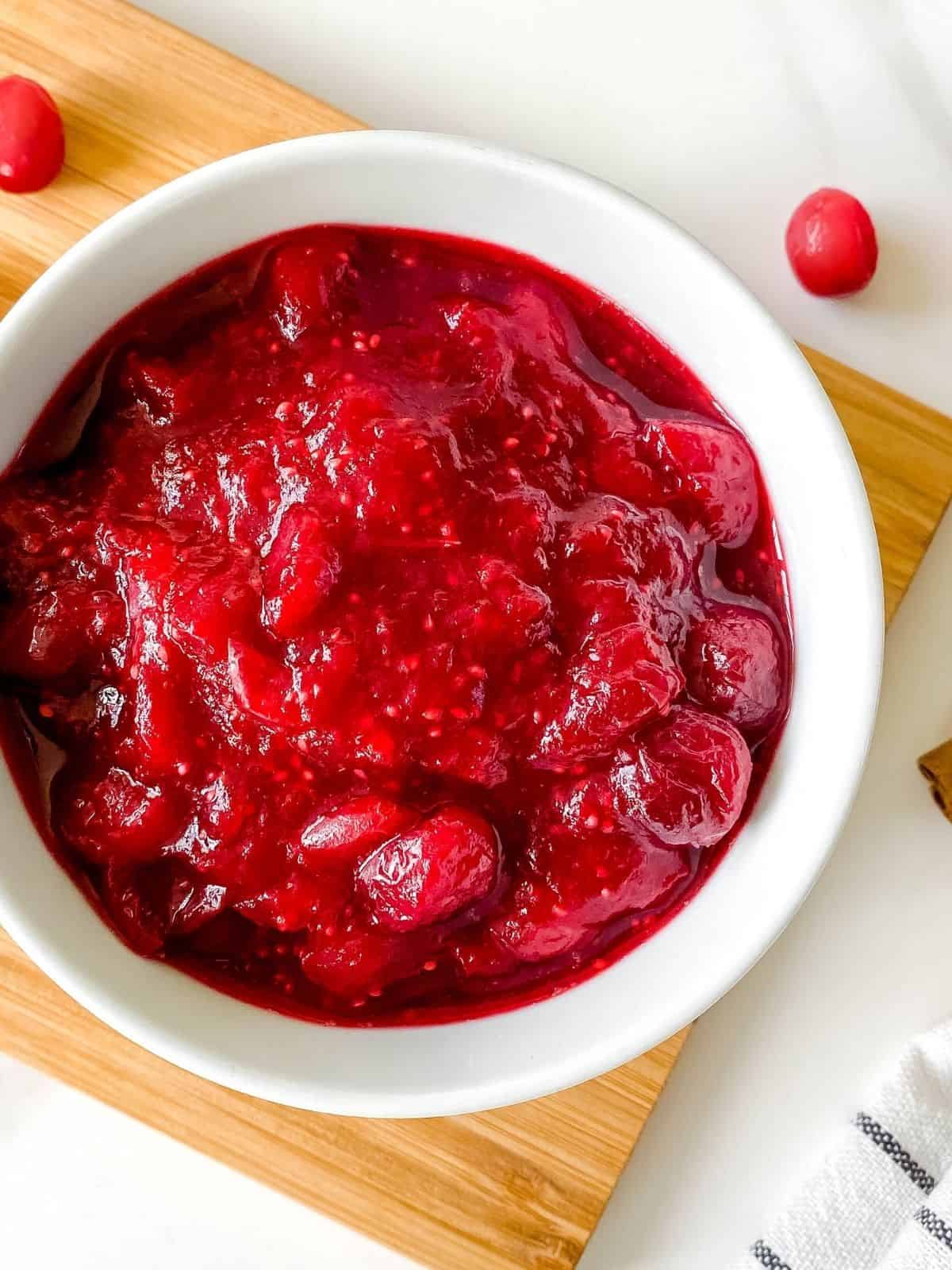 cranberry sauce in a white bowl on a wooden board next to a white and black striped cloth and fresh cranberries.