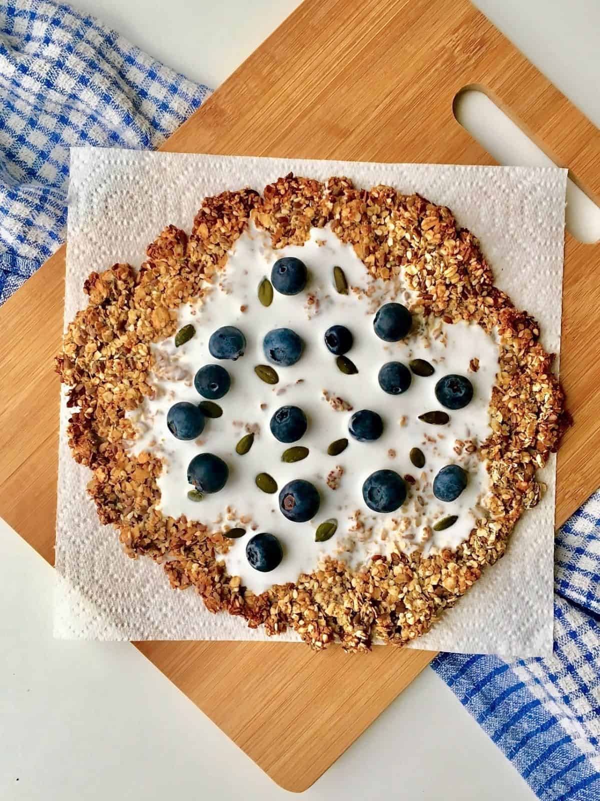 granola breakfast pizza on a wooden board on top of a blue checked tea towel.