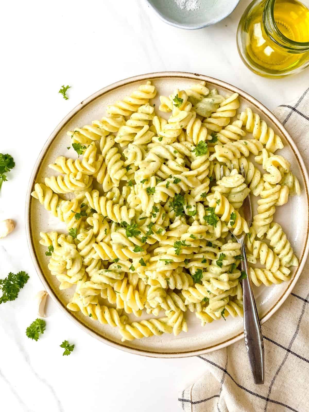 herbed tahini pasta on a brown plate on a checked tea towel next to a bottle of olive oil and bowl of salt.