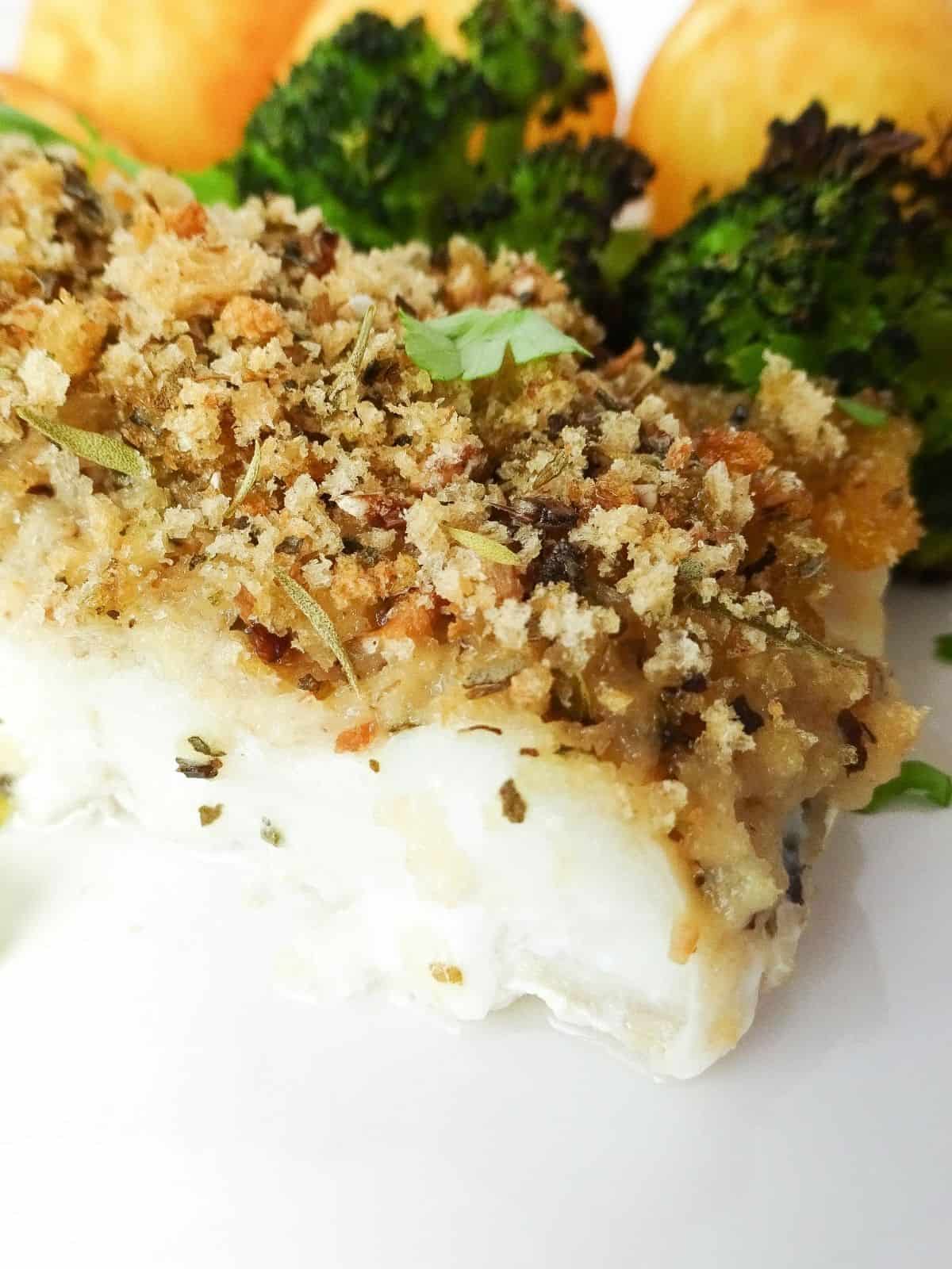 Italian herb crusted cod on a white plate with broccoli and potatoes next to it.