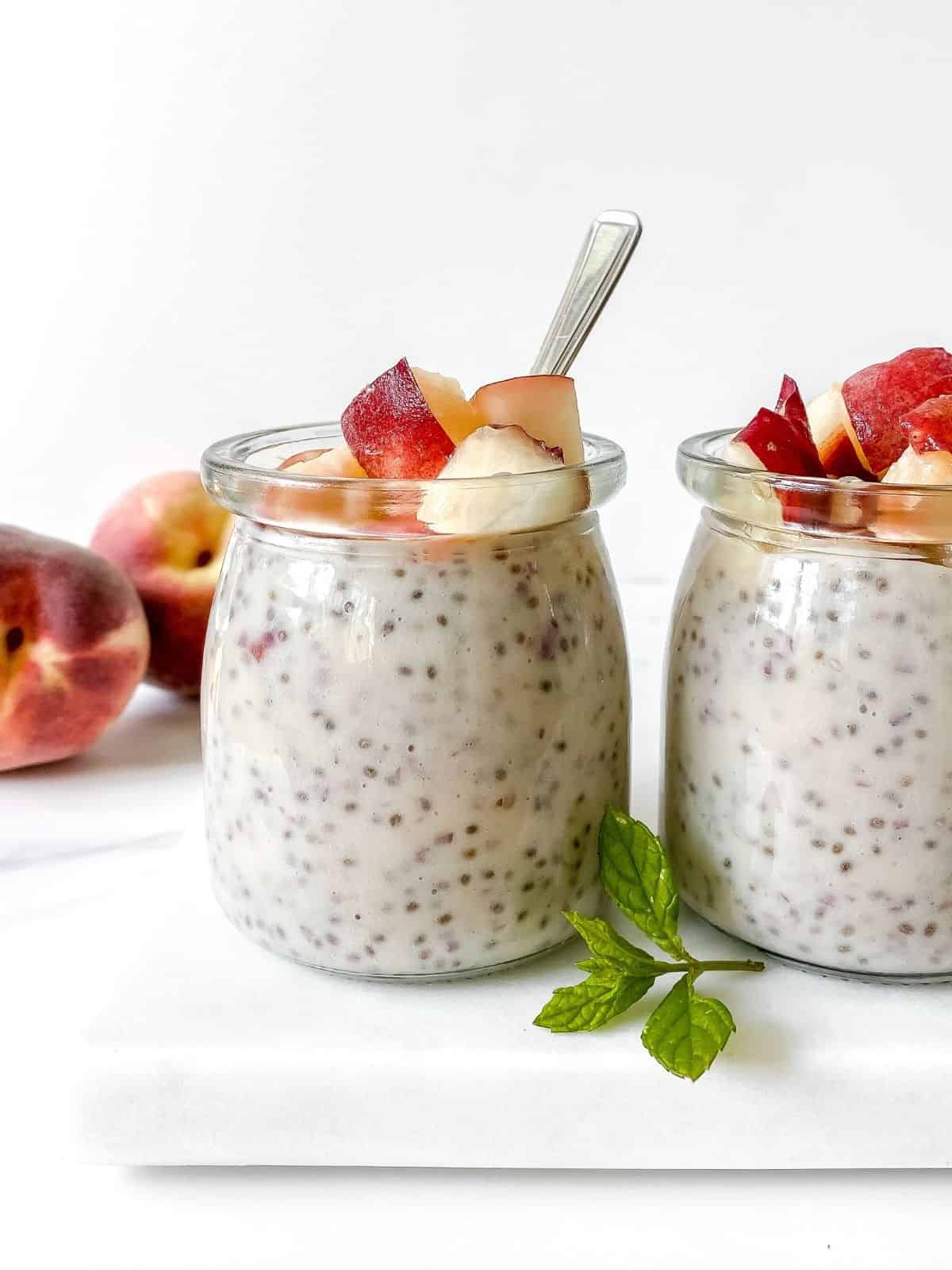 two glass jars of peach chia pudding with peaches in the background.