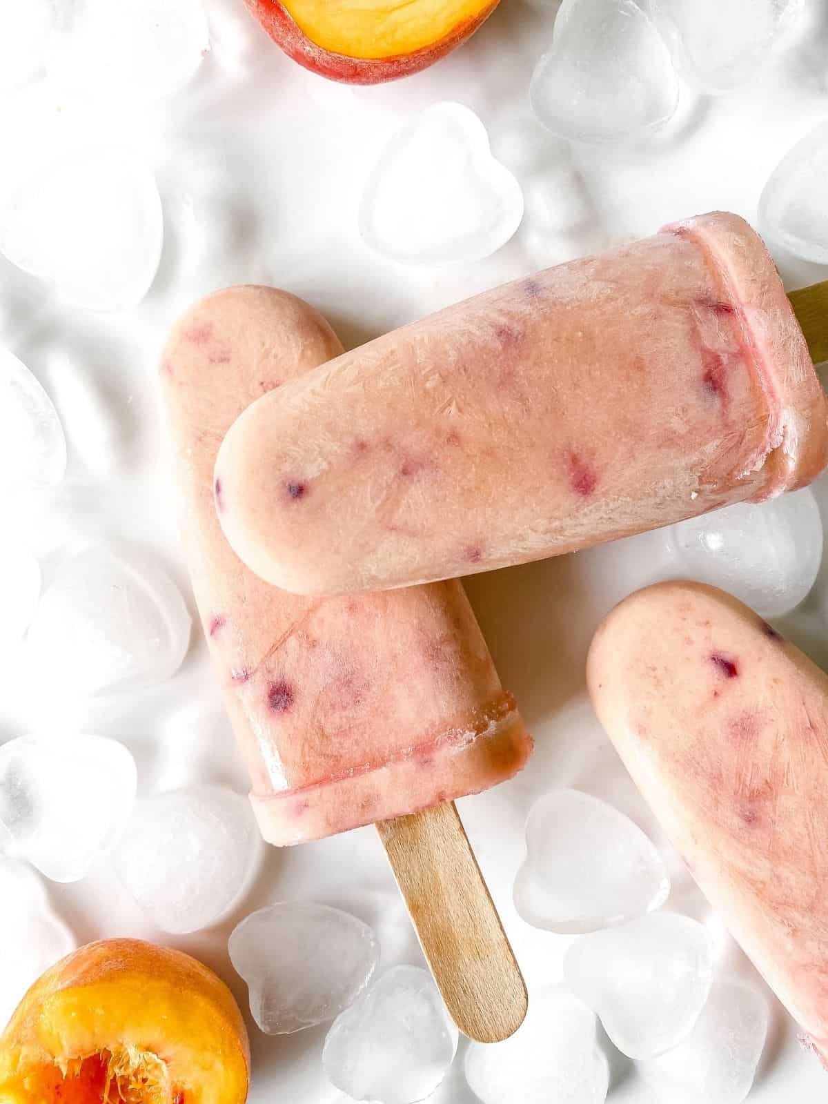 three peaches and cream popsicles surrounded by ice cubes and fresh peaches.
