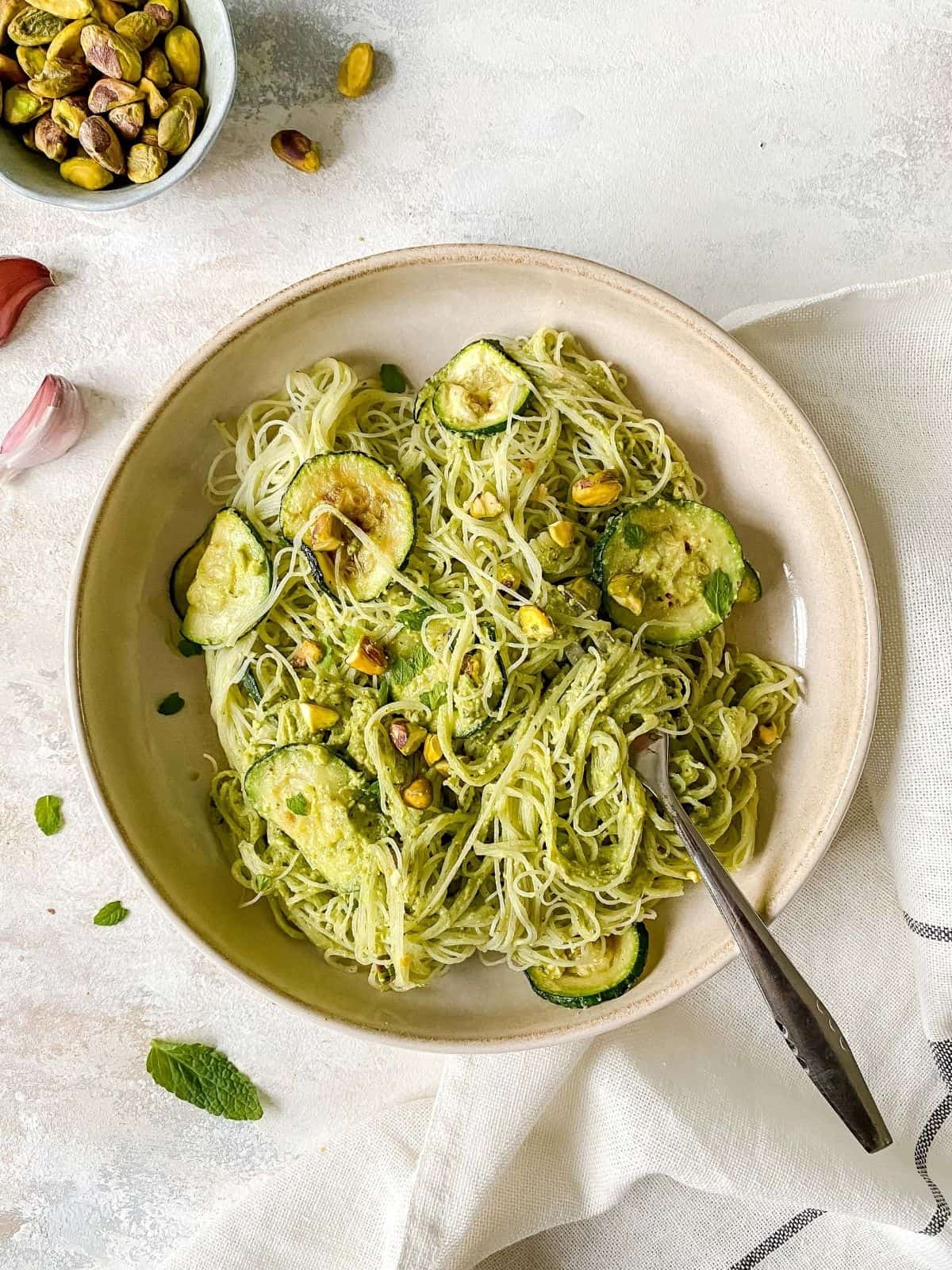 pistachio, zucchini and mint rice noodles in a brown bowl with a fork twisted into the noodles.