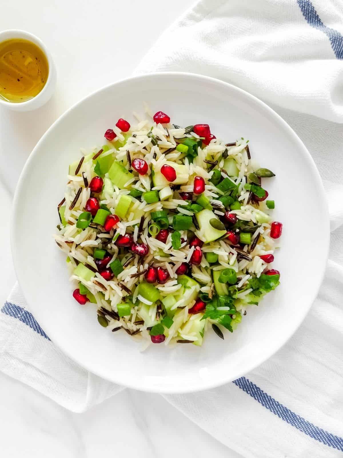 summer wild rice salad with pomegranate seeds on a white plate with a small bowl of dressing next to it.