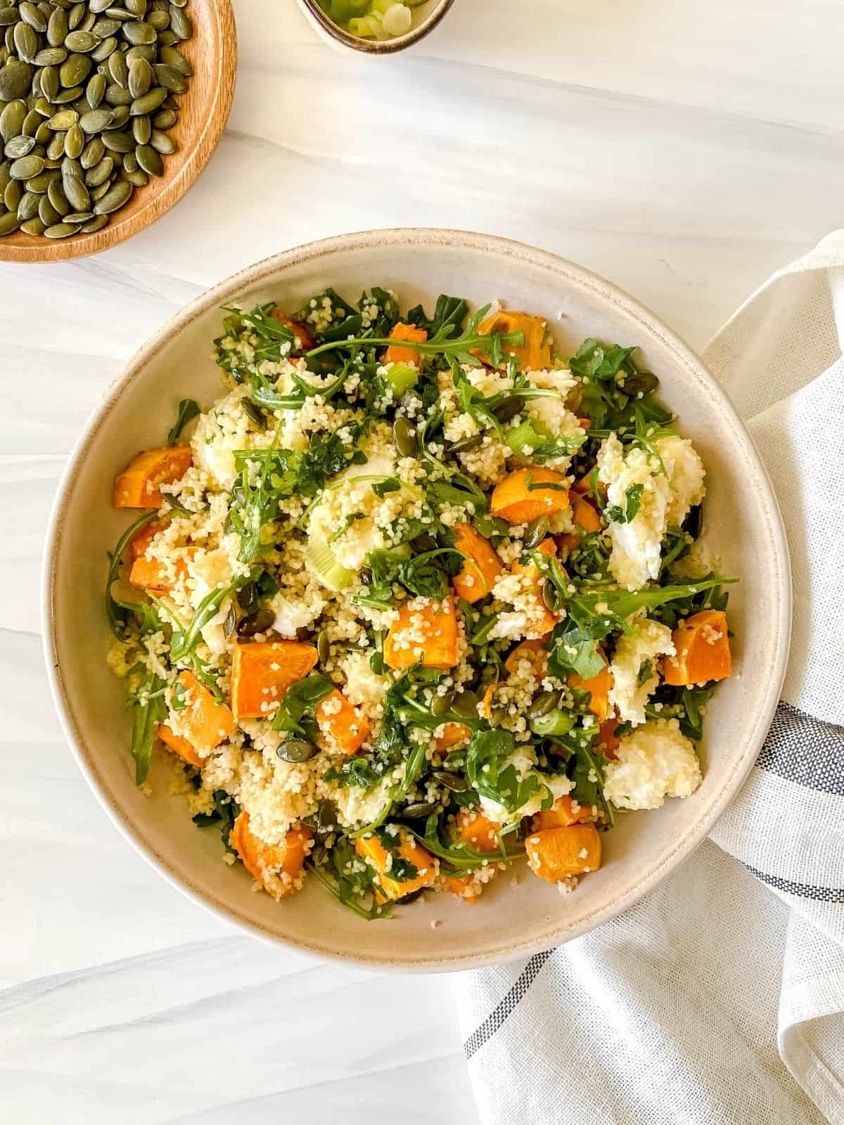 brown bowl of sweet potato couscous salad with small bowls of pumpkin seeds and green onion next to it.