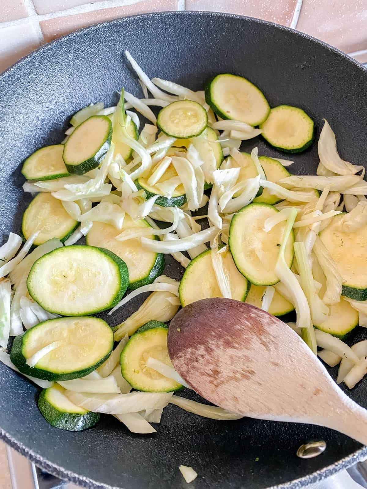 fennel and zucchini in a black skilelt being stirred with a wooden spoon.