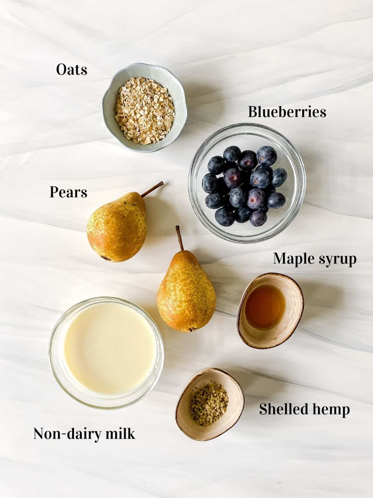 pears next to bowls of non-dairy milk, oats, maple syrup, shelled hemp and blueberries.