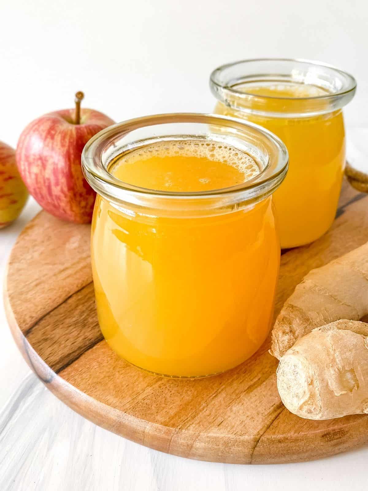 two glasses of apple ginger juice on a wooden board next to ginger root and apples.