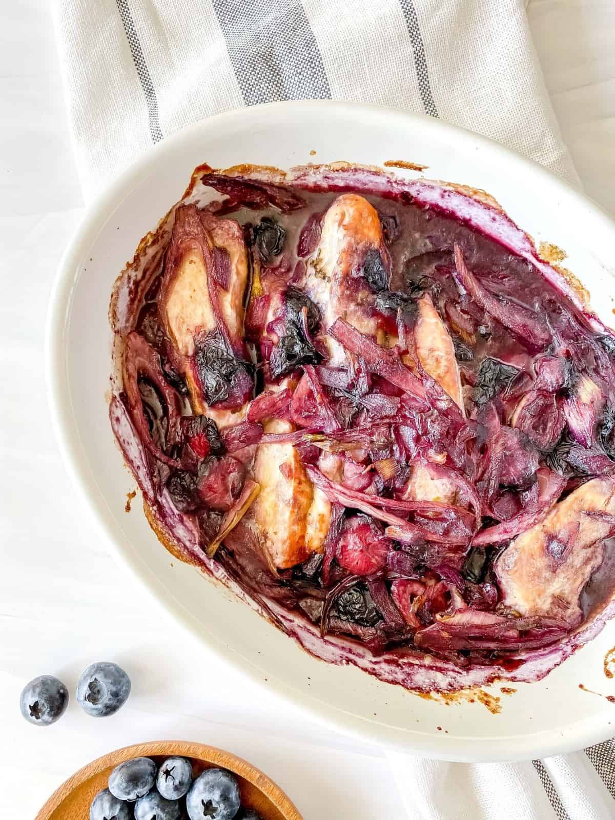baked blueberry chicken in a white ovenproof dish next to a small plate of blueberries.