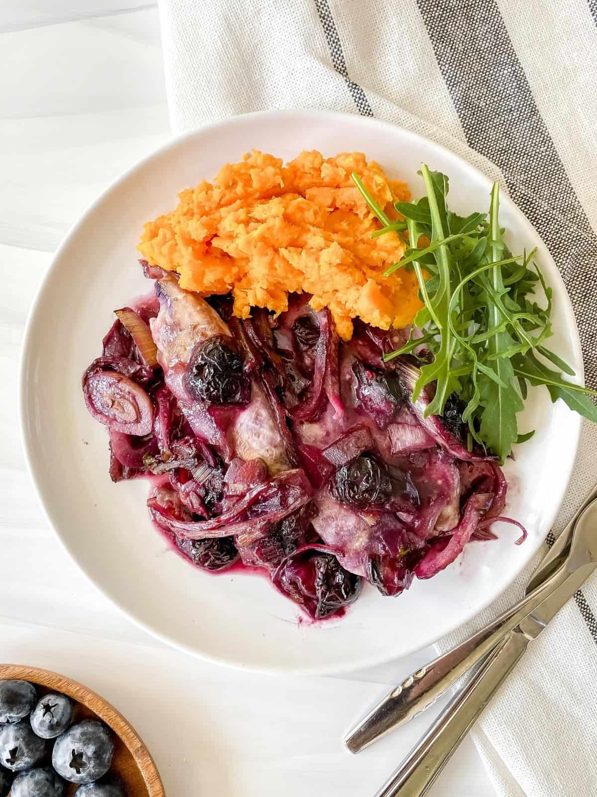 blueberry chicken, sweet potato mash and arugala on a white plate next to a brown plate of blueberries.