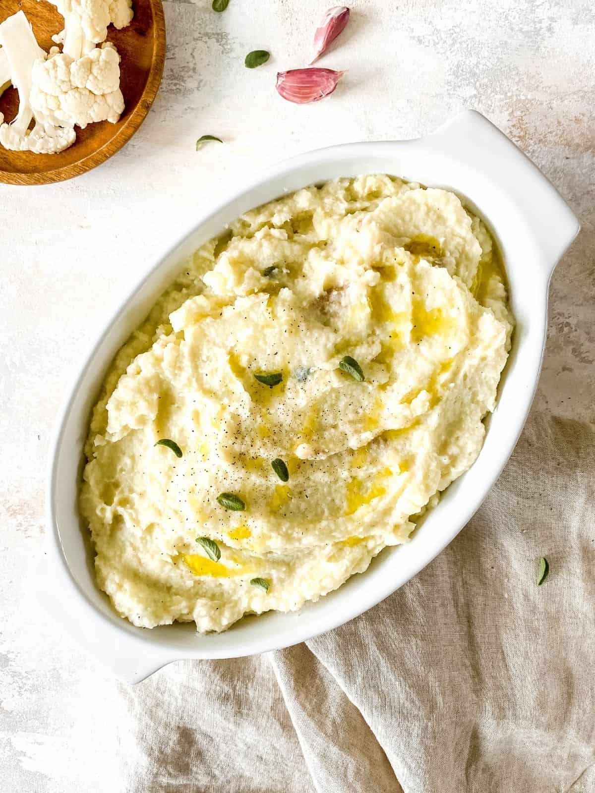 potato cauliflower mash in a white dish on a beige cloth with garlic next to it.