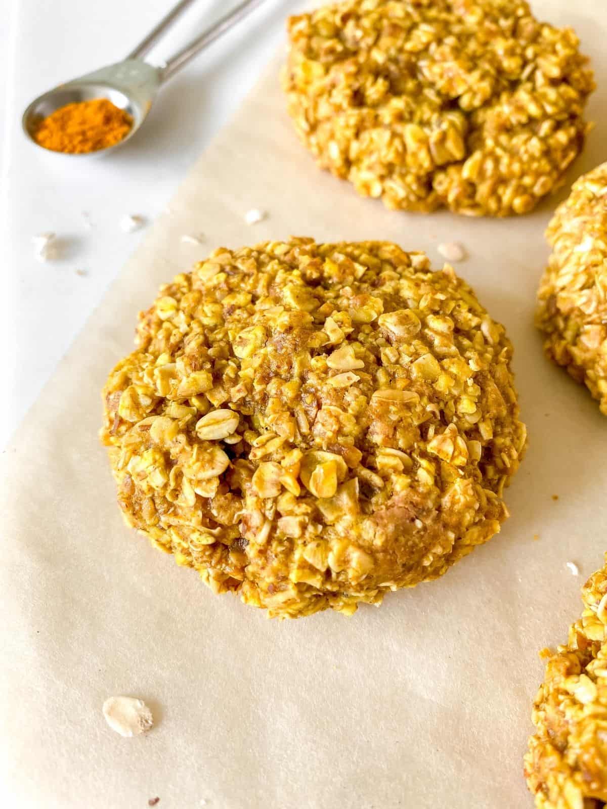 close up of a turmeric oatmeal cookies on parchment paper next to other cookies and a spoonful of turmeric.