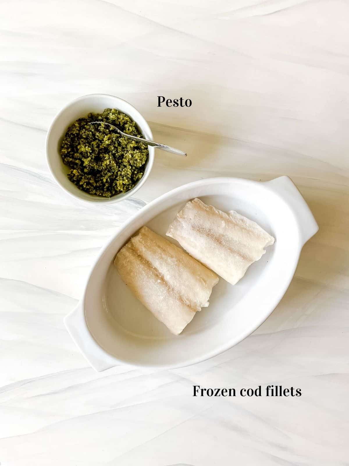 individually labelled two cod fillets in a white dish next to a white bowl of pesto.