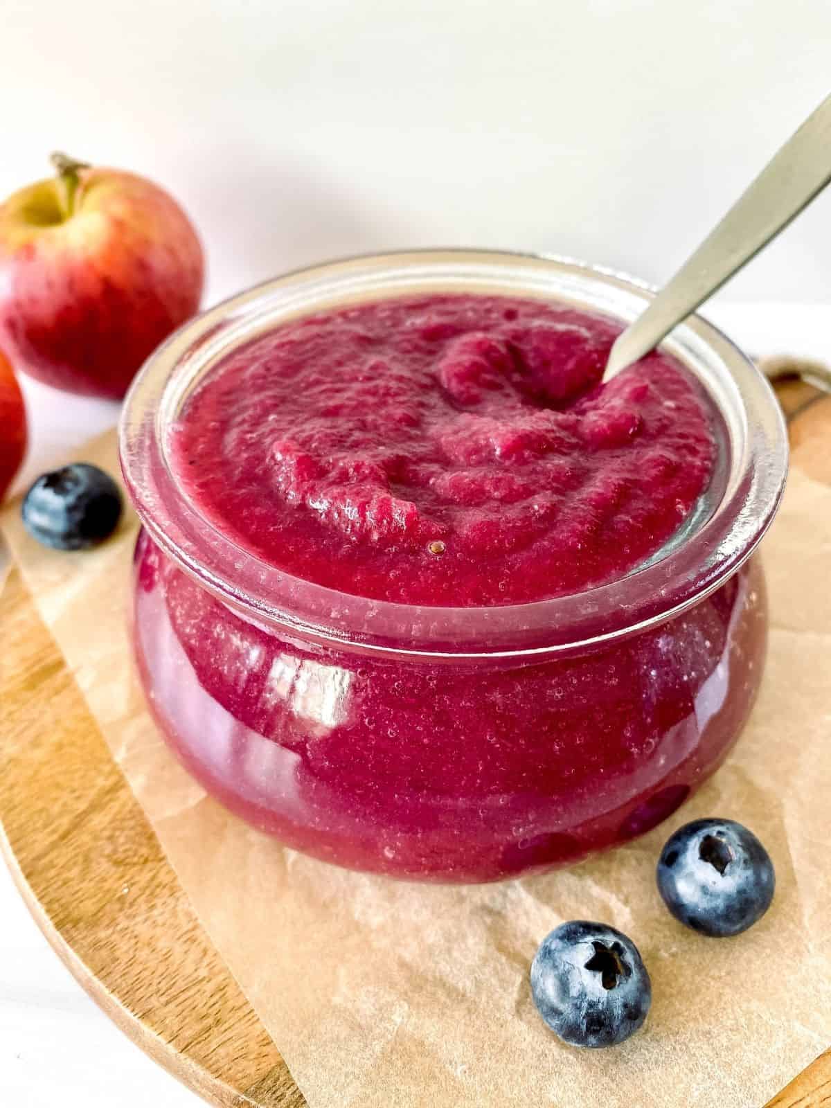 apple blueberry sauce in a glass jar with a spoon in it next to apples on a wooden board.