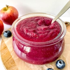 apple blueberry sauce in a glass jar on a wooden board with a spoon in it.