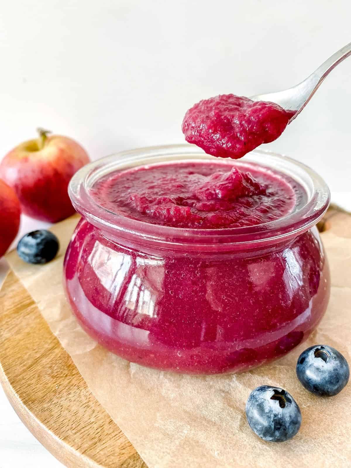 apple blueberry sauce in a glass jar with a spoonful of sauce above it and apples in the background.