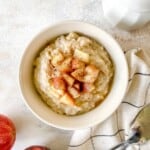 apple pie porridge in a cream bowl next to red apples and a white jug.