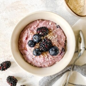 berry porridge in a cream bowl next to a spoon and blackberries.