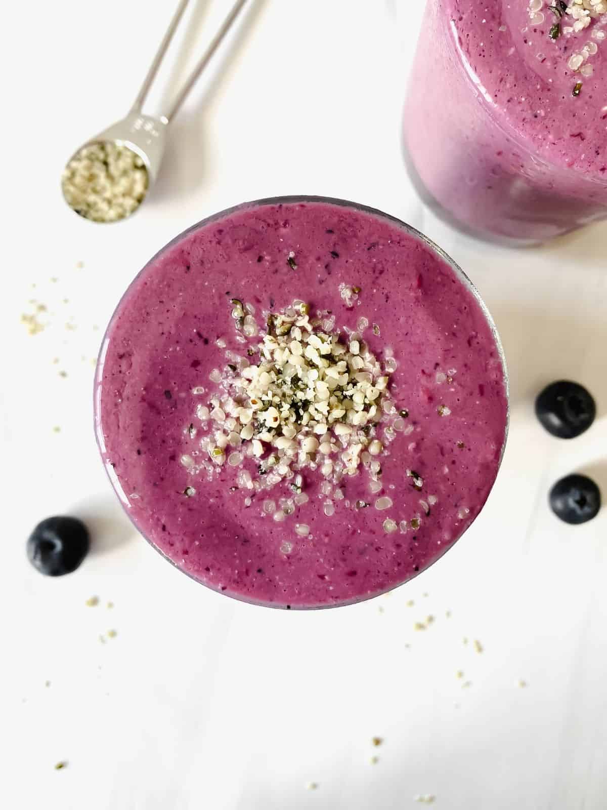 mixed berry smoothie in a glass next to a spoonful of hemp seeds and blueberries.