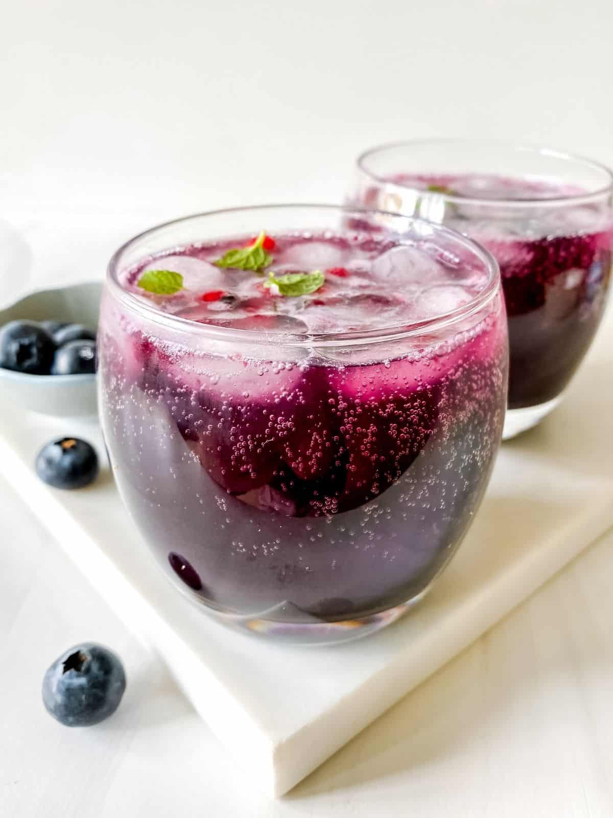 two glasses of blueberry mocktail with a blue bowl of blueberries in the background.