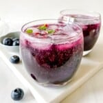 two glasses of blueberry mocktail with a blue bowl of blueberries in the background.