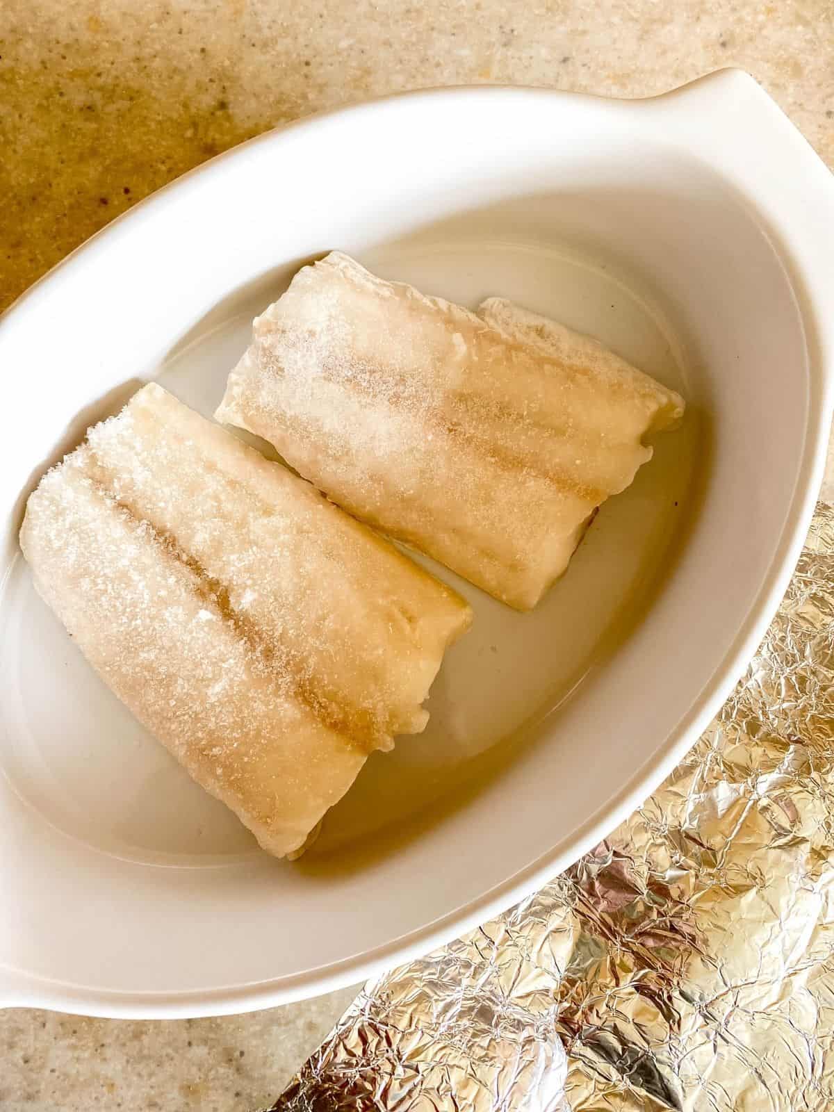 two cod fillets in a white dish next to foil.