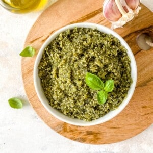 pumpkin seed pesto in a white bowl on a wooden board next to basil leaves and garlic bulb.