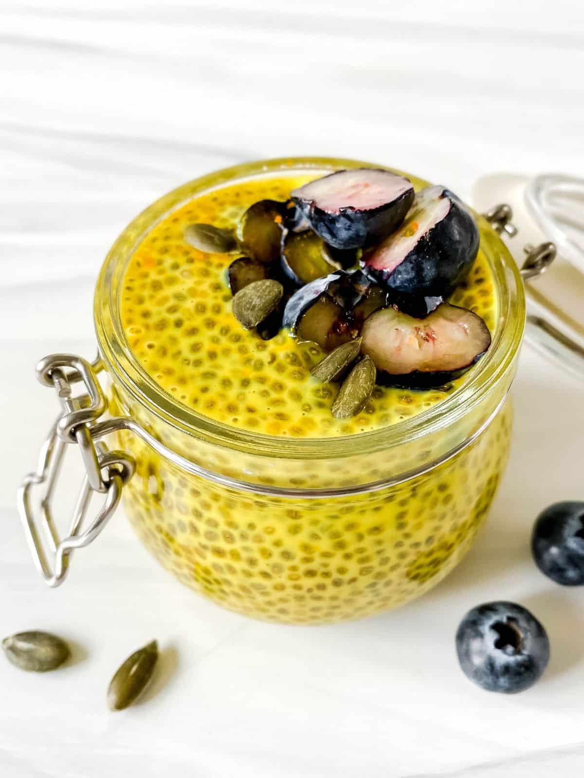 golden milk chia pudding in a glass jar with blueberries and pumpkin seeds next to it.